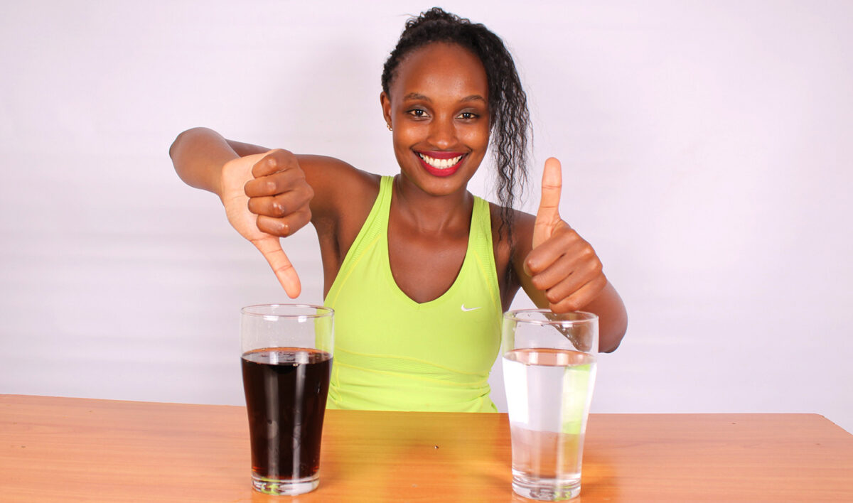 thumbs down for soda glass, thumbs up for water glass, pretty young black woman, broad smile, lime green exercise top, wooden table, off-white background